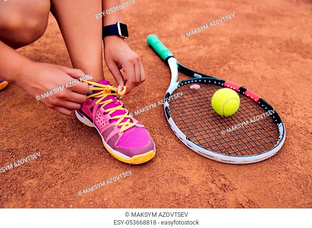 Athlete woman getting ready for playing a game of tennis, tying shoelaces. Close-up view of racket and ball