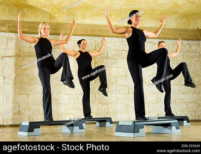 A group of women exercising in the fitness club. They're smiling and have upraised hands and one leg. Low angle view