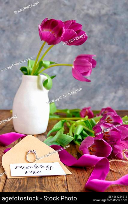 Bouquet of purple tulips in a vase, proposal ring and MARRY ME card over envelope on wooden table close up. Proposal scene with gemstone engagement ring