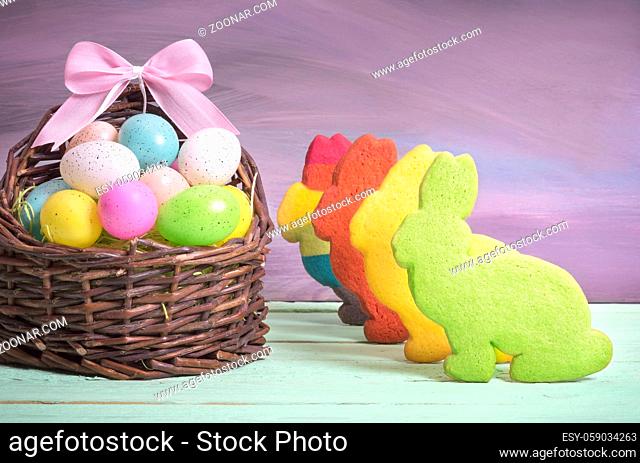 Easter card with a wicker basket full of painted eggs, with a pink ribbon bow and multicolor bunny shaped cookies, on a green table and purple wall