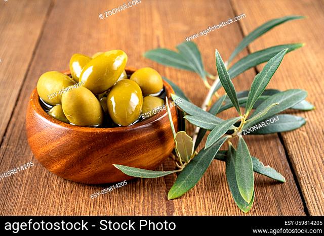 Green olives in olive oil with olive branch in a small wooden bowl on a rustic wooden background