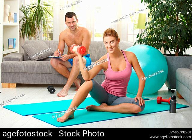 Young woman doing biceps exercise with dumbbell sitting on fittness mat at home, smiling