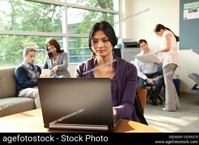 Wide shot of a woman sitting a table in busy breakroom working on laptop in office