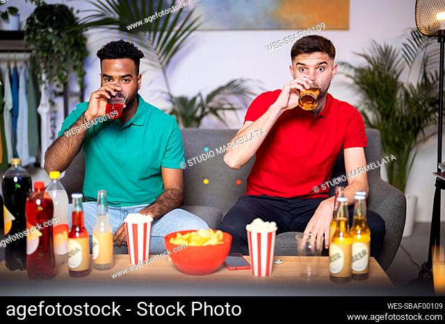 Young men drinking watching soccer match sitting on sofa in living room at home