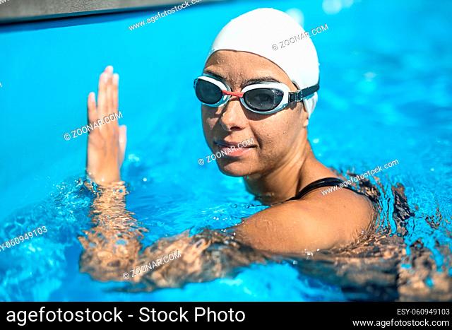 Amazing close-up photo of female swimmer in the swimming pool. She wears swimwear and swim glasses. Girl looks into the camera with a smile