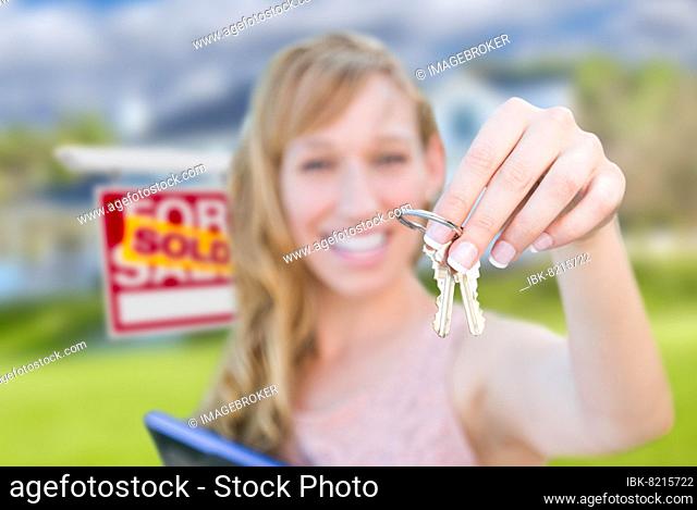 Excited woman holding house keys and sold real estate sign in front of nice new home