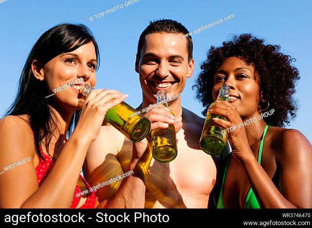 Group of friends - one man embraces two women and all have drinks in swimwear on the beach of a lake in summer