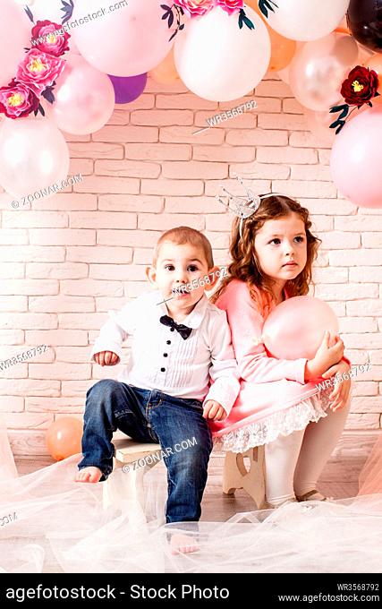 Boy and girl with crowns under birthday balloon and paper flower arch decorations. Childish photozone for celebration