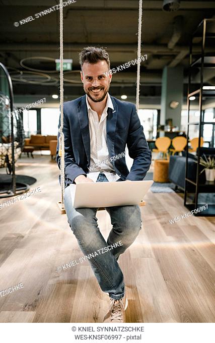 Businessman using laptop in coworking space, sitting on swing