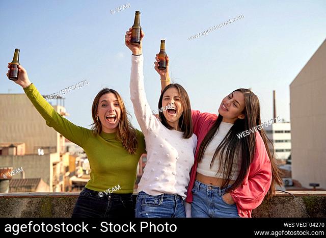 Happy female friends raising beer bottles enjoying on rooftop
