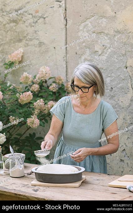 Mature woman sprinkling powdered sugar through sieve while preparing bread at backyard