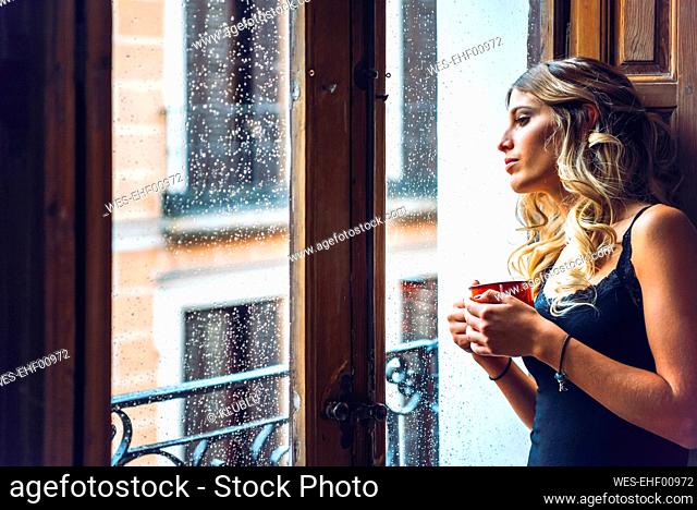 Beautiful young woman holding cup looking out of window