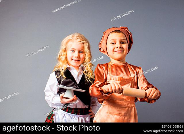 Two funny kids dressed in uniform are standing on the gray background. Boy chef and girl waiter