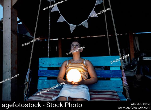 Girl holding illuminated lantern while sitting on swing at backyard