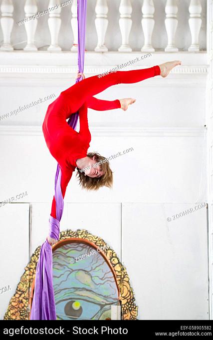 Flexible attractive woman air gymnast performs an exercise on aerial silk in a bright studio, telephoto shot