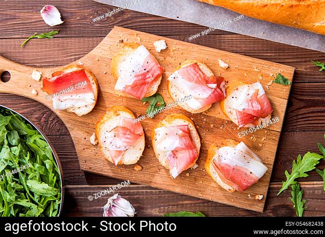 Photos baguette with bacon on wooden board on brown table with arugula