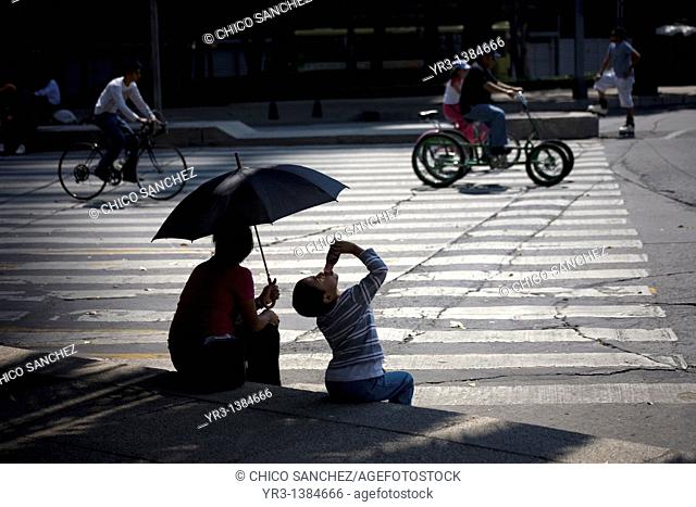 A boy drinks yogurt next to his mother holding an umbrella to shade them from the sun as they watch cyclists ride along Paseo de la Reforma Avenue in Mexico...