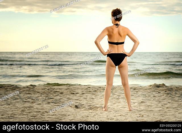 beautiful woman in a black swimsuit is looking at the sea on the beach at sunset