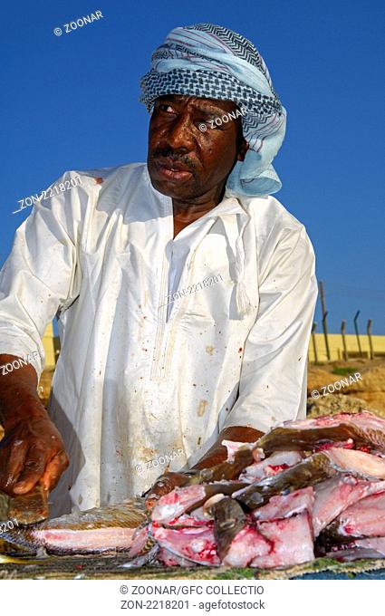 Dunkelhäutiger Fischhändler beim Zerkleinern von frischem Fisch an einem Verkaufsstand im Freien, Sur, Sultanat Oman / Dark-skinned fishmonger cutting fresh...