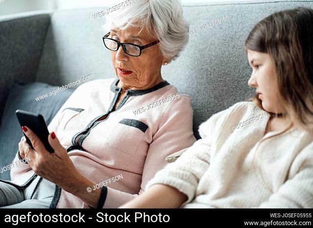 Grandmother using mobile phone with granddaughter on sofa