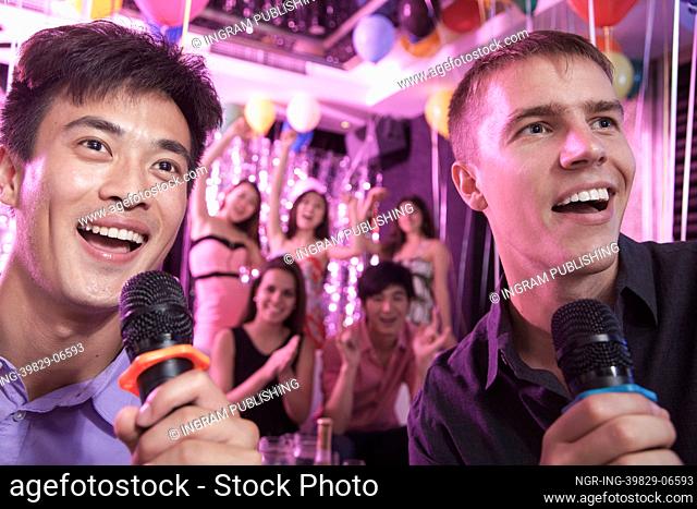 Two friends holding microphones and singing together at karaoke, friends in the background