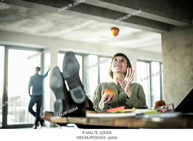 Woman juggling apples in the office, sitting with feet on desk