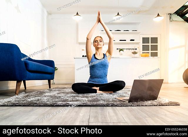 White caucasian fit sporty woman doing yoga Sukhasana sit pose in living room at home watching online tutorials from trainer on laptop