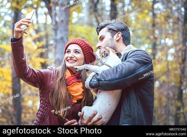 Woman and man with their dog on autumn walk taking a phone selfie posting it online on social media
