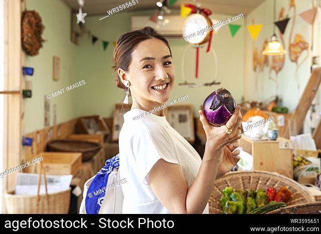 Two smiling Japanese women standing in a farm shop, holding aubergine, looking at camera