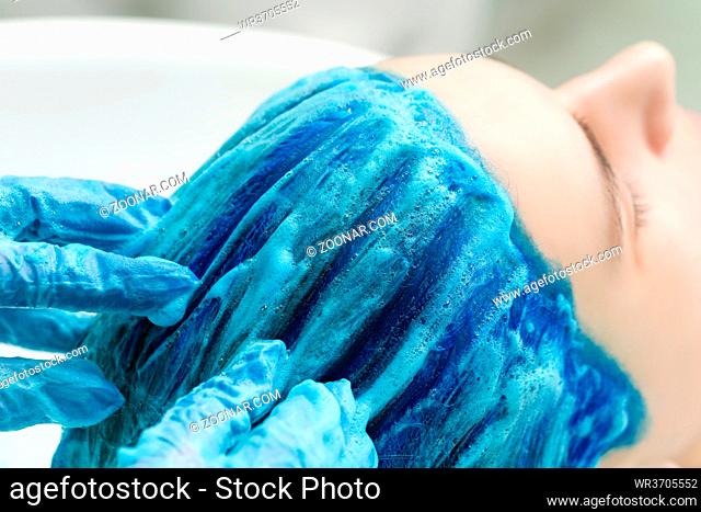 Professional hairstylist wearing protective gloves washes client's sapphire color hair with conditioner shampoo for washing after process of coloring hair in...