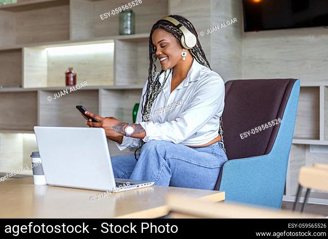 African American woman wearing headphones using cellphone in home with a laptop in front of her. Concept of people in home