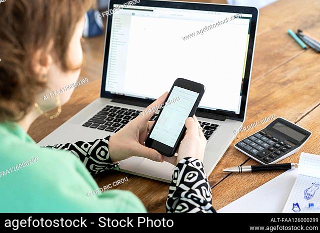 Woman using smartphone while working on laptop at home
