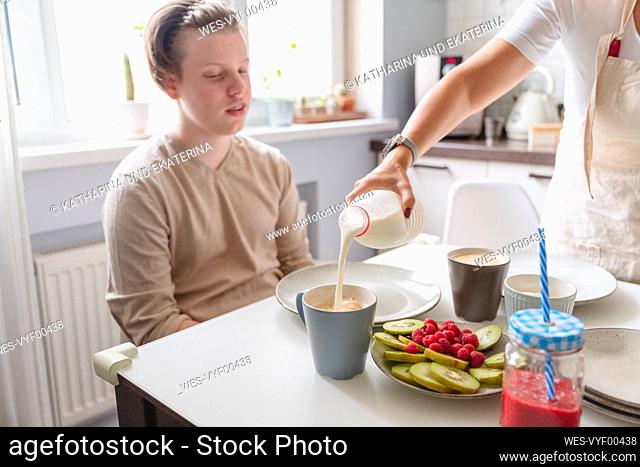 Woman pouring milk in cup for teenage son at dining table in kitchen