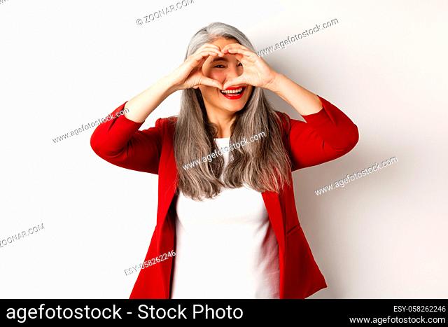 Lovely asian middle-aged woman with grey hair, wearing red blazer, showing heart sign and peeking throught it, I love you gesture
