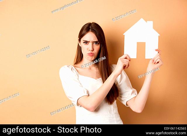 Real estate. Angry girl showing house cutout and frowning upset, complaining on apartment, standing against beige background