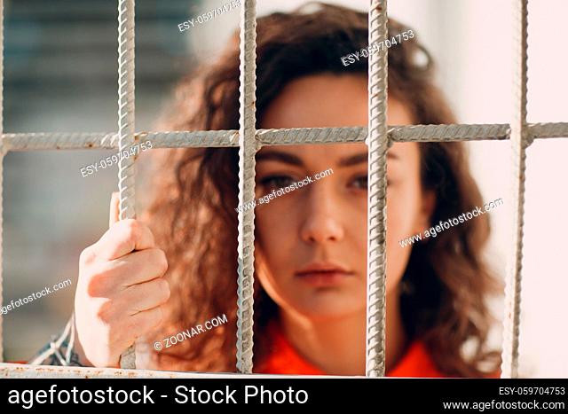 Young brunette curly woman in orange suit behind jail bars. Female in colorful overalls portrait