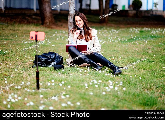 smiling female student learning remotely outdoors in park. Girl online learning courses using smartphone sitting in park