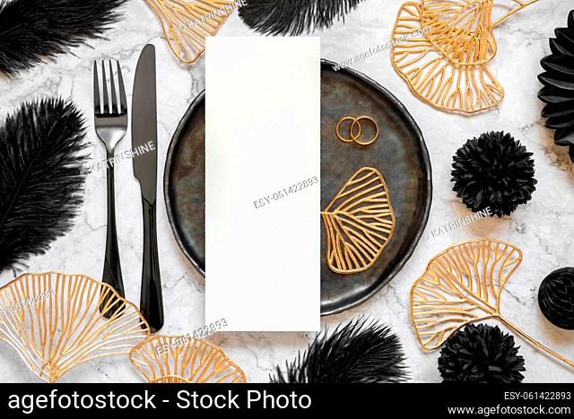 Black and golden wedding table setting with a blank card and rings top view, mockup. Elegant flat lay with vertical menu or Invitation paper card