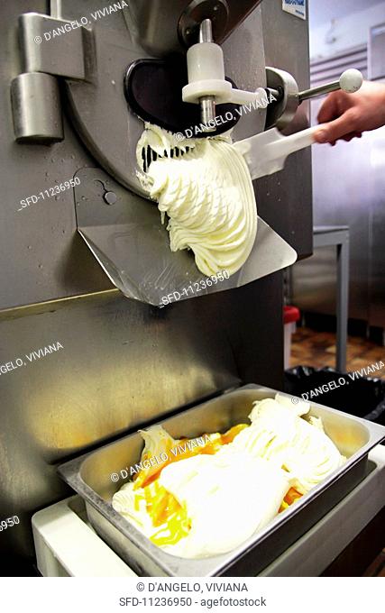 An ice cream machine producing a pale ice-cream mixture, a hand helping to release the ice cream with a spatula