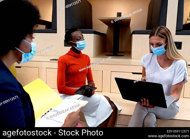 Diverse group of female business colleagues wearing masks holding documents using tablet and laptop