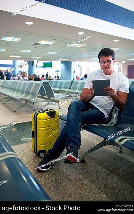 Young man sitting in the airport lounge and using tablet computer, yellow suitcase nearby. Easy work or entertainment on travel