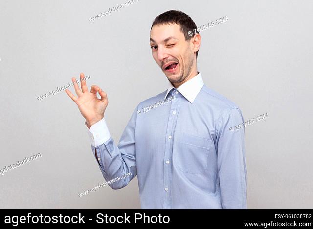 Portrait of funny crazy handsome bristle businessman in classic light blue shirt standing with Ok sign and looking at camera, winking