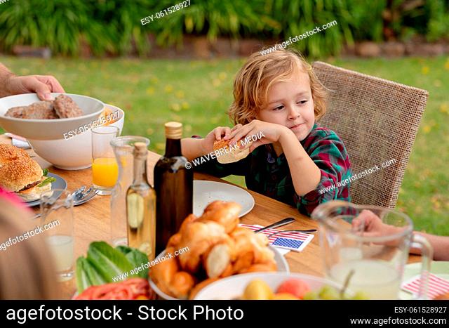 Smiling caucasian boy holding hamburger eating meal with family in garden