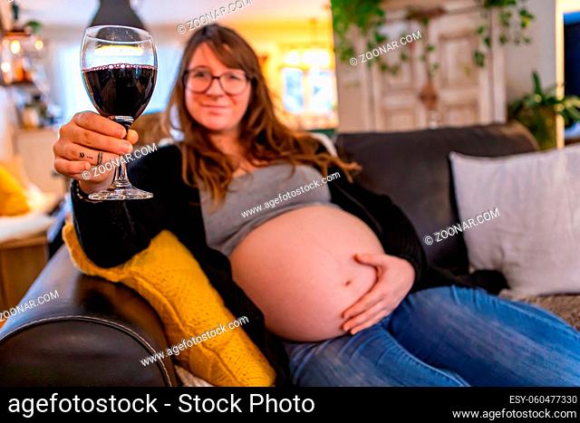 A shallow depth of field as a blurry pregnant woman relaxes on sofa in family room with glass of red wine, risking fetal alcohol syndrome