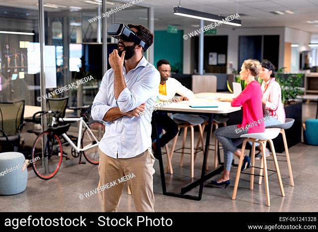 Thoughtful caucasian businessman wearing virtual reality simulator against biracial colleagues