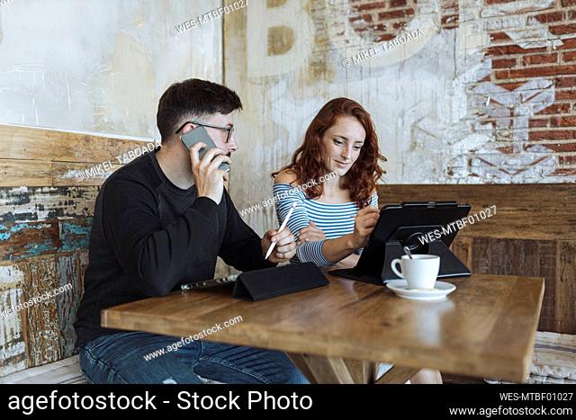 Male professional talking on smart phone while sitting with female colleague working at cafe