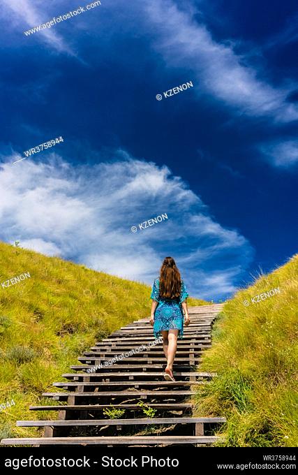 Full length rear view of a young woman wearing a blue dress while climbing wooden stairs outdoors in an idyllic travel destination from Indonesia