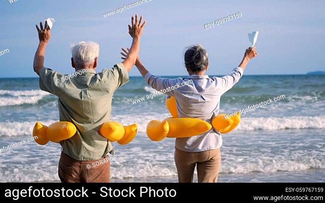 Happy senior couple wear rubber ring take off medical face mask and running to the beach enjoying life, relaxing after win coronavirus pandemic (COVID-19)