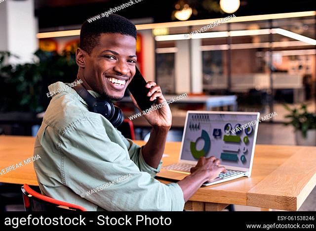 Portrait of smiling african american businessman talking on phone while planning strategy on laptop