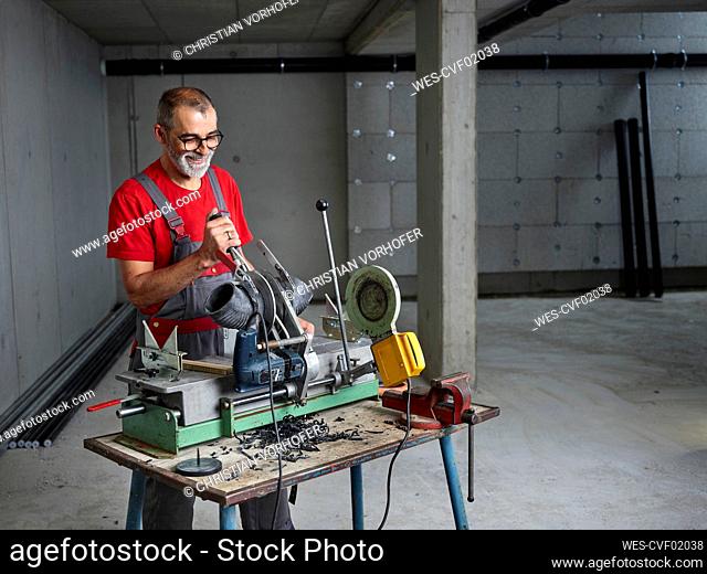 Smiling plumber wearing eyeglasses cutting plastic pipe at workbench